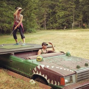 Two golfers posing with the hole nine teeing area of the Decatur Nine. An old, rusty island vehicle serves as the platform for teeing off.